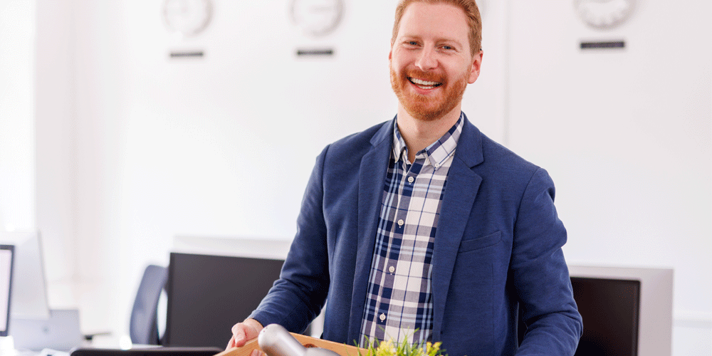 Man in blue suit jacket and button-down shirt as he holds a packed box of office possessions.
